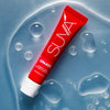 Eine rote Tube Opakes Cosmetic Paint liegt auf einer nassen blauen Oberfläche mit Wassertropfen.