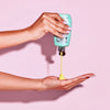Rapid Tanning Elixir aus einer grünen Flasche wird auf eine Hand vor rosa Hintergrund gegeben.