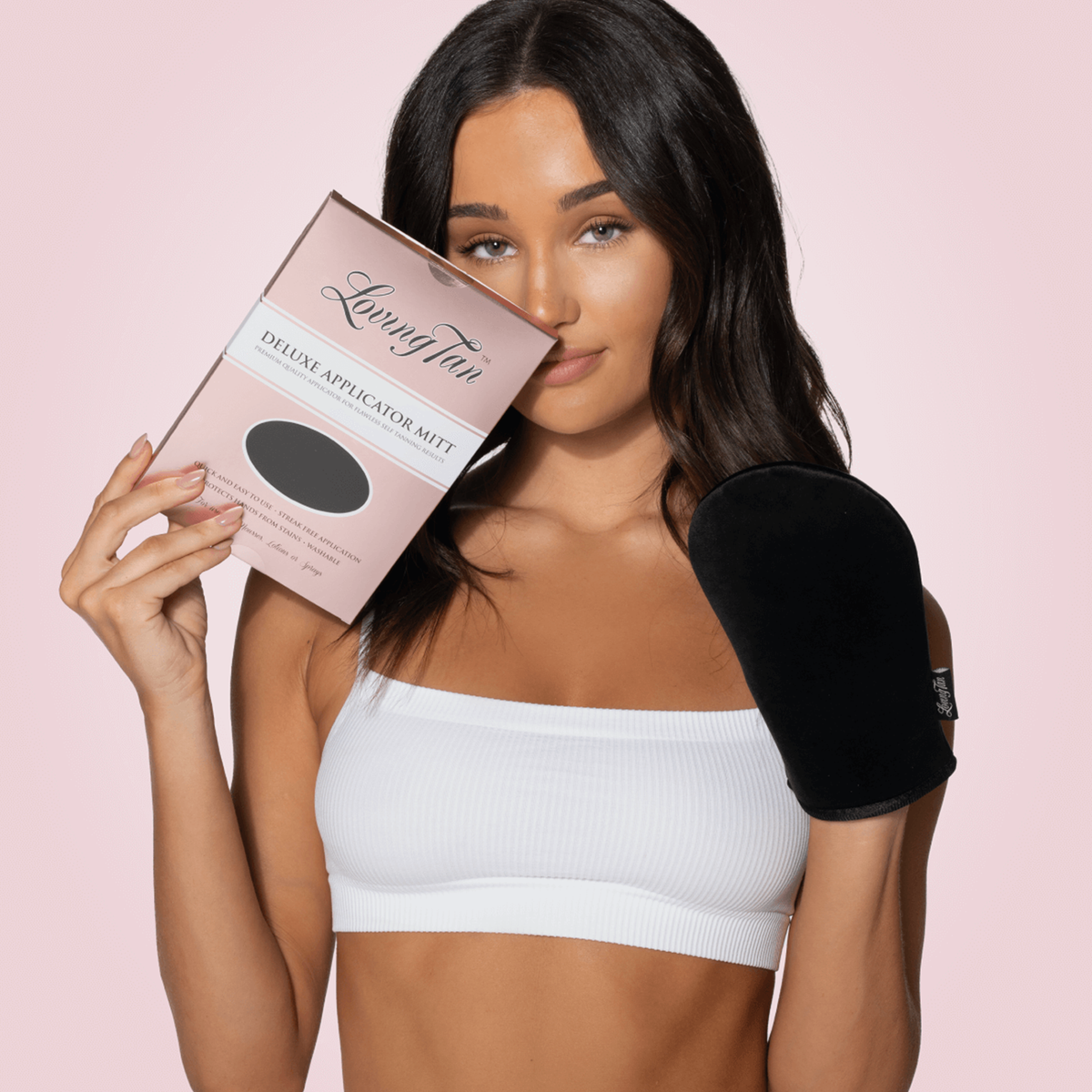 Frau mit weißem Top hält eine Verpackung des Loving Tan Deluxe Applicator Mitts in der einen Hand und trägt den schwarzen Bräunungshandschuh an der anderen.