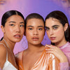 Drei Frauen deren Lippen mit dem Gloss Matrix Lipgloss von Mesauda Beauty geschminkt sind posieren nebeneinander vor einem lilanen Hintergrund.