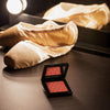 Das Mesauda Beauty At First Blush Rouge liegt auf einer dunkles Ablage, dahinter liegen Balettschuhe. 