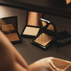 Verschiedene Töne des Mesauda Beauty Nude Venus Puders halbgeöffnet und verteilt auf einer dunklen Ablage. Ein Produkt geschlossen.

