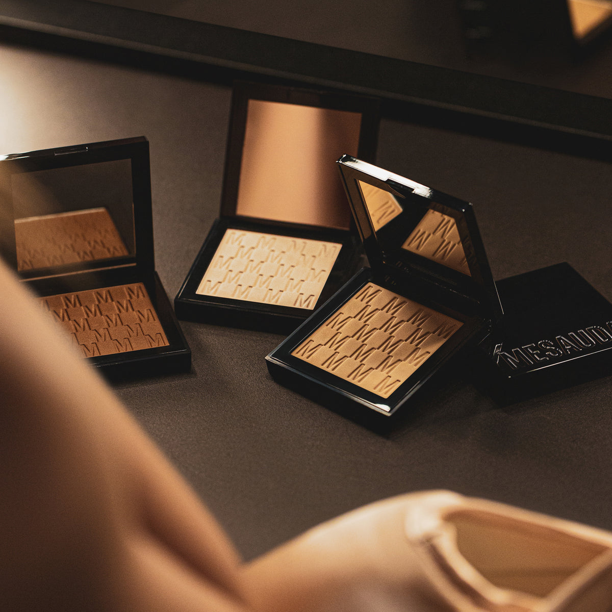 Verschiedene Töne des Mesauda Beauty Nude Venus Puders halbgeöffnet und verteilt auf einer dunklen Ablage. Ein Produkt geschlossen.
