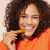 Lächelnde Frau mit lockigem Haar hält eine NUXE Honey Lip Care Rêve de Miel vor ihr Gesicht und trägt ein orangefarbenes Hemd.