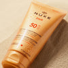Eine orangefarbene Tube NUXE Sun Delicious Lotion High Protection SPF 50 mit Wassertropfen liegt auf sandigem Boden.