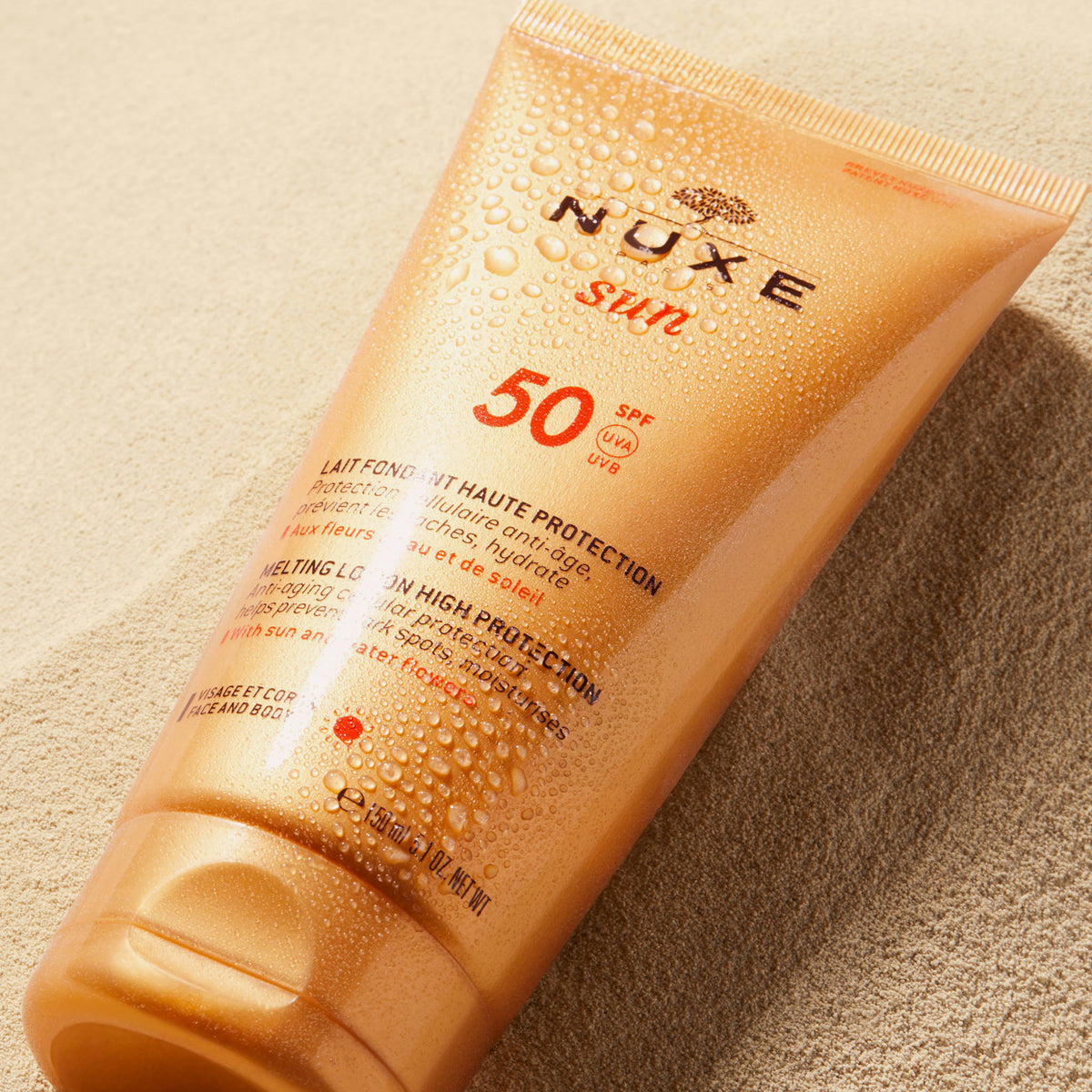 Eine orangefarbene Tube NUXE Sun Delicious Lotion High Protection SPF 50 mit Wassertropfen liegt auf sandigem Boden.