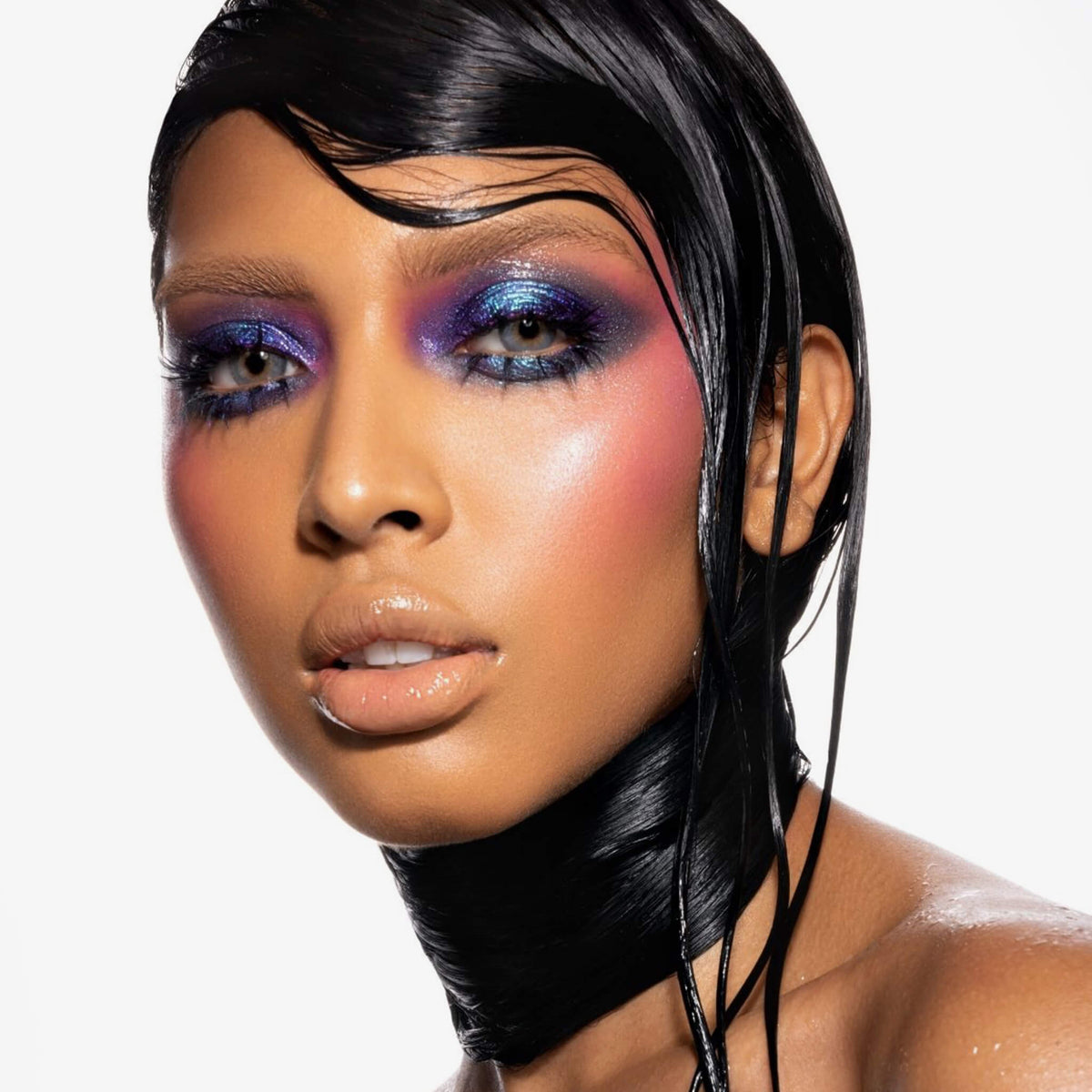 Eine Frau trägt kräftiges lilafarbenes Twin Flames-Augen-Make-up, glänzende Lippen und hat ihr nasses schwarzes Haar um den Hals gewickelt.