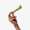 Eine Hand hält eine goldene Tube LH Cosmetics Shaping Light Warm Glow vor schlichtem weißen Hintergrund.