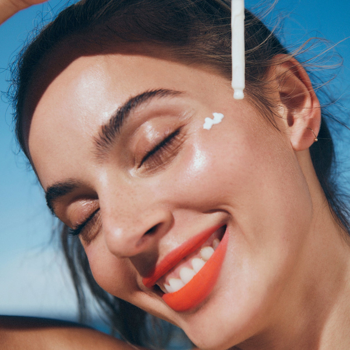 Eine Frau mit leuchtendem Lippenstift lächelt, während sie vor dem Hintergrund eines blauen Himmels Queen Screen™ SPF50+ in der Nähe ihres Auges aufträgt.