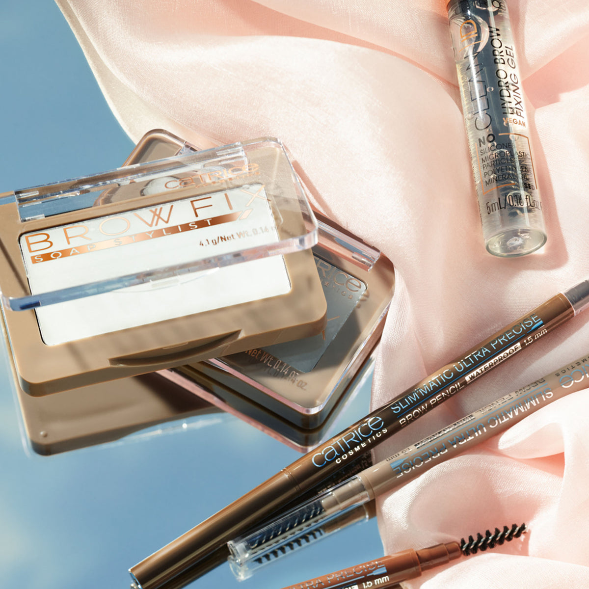 Die Brow Fix Soap Stylist, zusammen mit Stiften und Serum, präsentiert auf einem zartrosa Stoff.