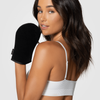 Eine Frau in einem weißen Oberteil hält den schwarzen Loving Tan Deluxe Self Tanning Applicator Mitt und blickt über ihre Schulter in die Kamera.