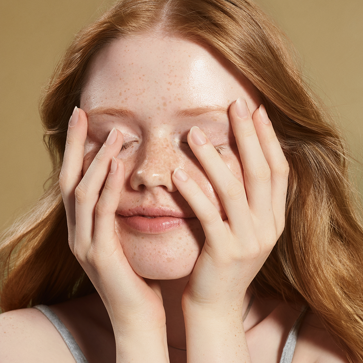Eine Person mit langen roten Haaren lächelt sanft und bedeckt ihre Augen mit den Händen, während sie nach der Verwendung der Advanced Snail 96 Mucin Power Essence vor einem beigen Hintergrund strahlende Haut zur Schau stellt.