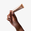 Eine Hand hält eine bronze-goldene Tube LH Cosmetics Shaping Light Desert Glow vor schlichtem weißen Hintergrund.