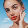 Mit leuchtend orangefarbenem Lippenstift und Queen Screen™ Luminising Skinscreen™ SPF50+ berührt eine Person vor dem Hintergrund eines blauen Himmels ihre Wange.