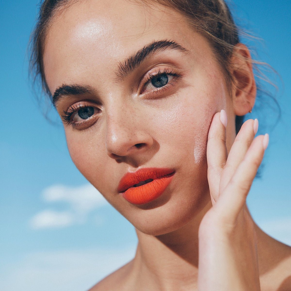 Mit leuchtend orangefarbenem Lippenstift und Queen Screen™ Luminising Skinscreen™ SPF50+ berührt eine Person vor dem Hintergrund eines blauen Himmels ihre Wange.